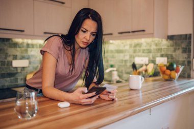 Orta yaşlı bir kadın elinde banka kartı tutuyor ve evinde bir mutfakta dururken mobil bir uygulamayla online alışveriş yapmaktan zevk alıyor. Çevrimiçi satın alma kavramı.