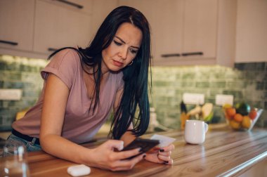 Güzel orta yaşlı kadın mutfakta duruyor ve akıllı telefonundan kredi kartıyla online ödeme yapıyor. Çevrimiçi alışveriş, para harcamak, hayatın tadını çıkarmak..