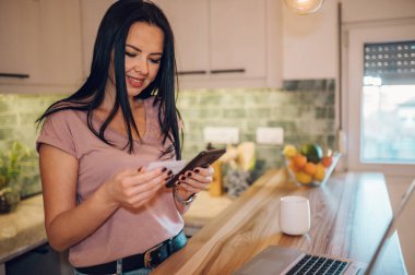 Orta yaşlı bir kadın elinde banka kartı tutuyor ve evinde bir mutfakta dururken mobil bir uygulamayla online alışveriş yapmaktan zevk alıyor. Çevrimiçi satın alma kavramı.