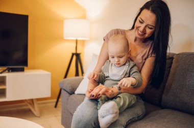 Küçük oğluyla kanepede vakit geçiren mutlu genç bir anne. Aile, bebek ve annelik kavramı. Anne oğul kaynaşıyor..