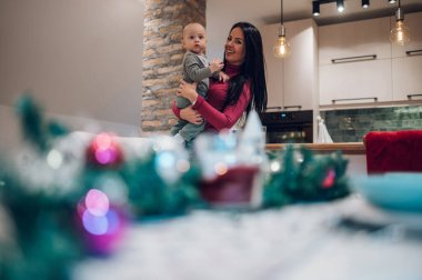 Anne, Noel süslemeli bir odada durup eğlenirken bebeğini kucağına alıyor. Annem kucağında bir bebekle Noel arifesini kutluyor. Noel ve Yeni Yıl konsepti.