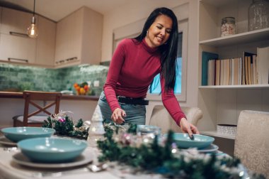 Mutlu orta yaşlı anne ailesiyle Noel yemeği için evde masayı hazırlıyor. Kadın ailesiyle tatilini geçirmek için tabakları servis ediyor ve dekore ediyor. Boşluğu kopyala.