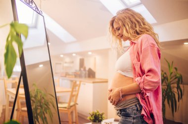 Portrait of a happy young pregnant woman tenderly touching her belly at home and looking herself in the mirror. Beautiful awaiting mother embracing her tummy and smiling. Enjoying pregnancy time. clipart