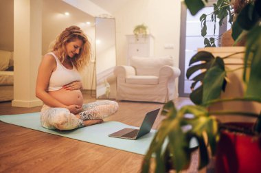 Genç hamile bir kadın rahat bir oturma odasında yoga pozisyonunda çalışırken dizüstü bilgisayarda doğum öncesi dersi izliyor. Sağlıklı Hamilelik Yogası ve Spor Konsepti. İki koluyla karnını tutuyor.