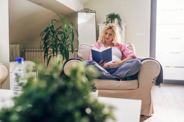 Mutlu hamile kadın evde bir sandalyede oturup karnına dokunarak kitap okuyor. Hamilelik, annelik, insanlar ve beklenti konsepti. Boşluğu kopyala.