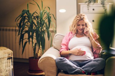 Evdeki bir oturma odasında rahat bir sandalyede dinlenirken akıllı telefon kullanan, gülümseyen Kafkasyalı hamile kadın ve gelecek olan bir çocuk için internetten hazırlıklar yapıyor. Cep telefonuyla konuşuyorum. Boşluğu kopyala.