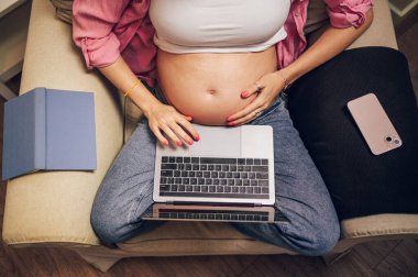 Evde dizüstü bilgisayar kullanan hamile bir kadının en iyi görüntüsü. Müstakbel anne sandalyede otururken bilgisayar kullanıyor. Arkadaşlarla ve ailelerle iletişim, online alışveriş ya da hamilelik sırasında iş.