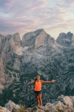 Dağın tepesinde dikilen mutlu bir kadın gökyüzü koşucusu özgürlüğünü yaşıyor. Arka planda dağ zirvesi ve bulutlu sabah gökyüzü var. Bir sporcu kadın gökyüzünde koşmaktan zevk alıyor..