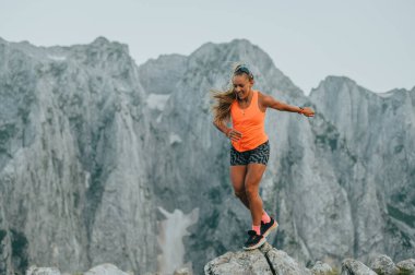 Aşırı sporcu bir kadın dağ sırasındaki kayalık arazide koşuyor ve zıplıyor. Bir atlet dağlarda vahşi doğada koşuyor. Bir skyrunner zıplıyor ve koşuyor. Arka planda dağ sırası var..