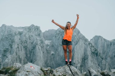 Aşırı sporcu bir kadın dağ yamacındaki bir taşın üzerinde duruyor ve başarısını kutluyor. Başardığı bir amacı kutlayan bir skyrunner. Mutlu bir sporcu dağlarda vahşi doğada duruyor..