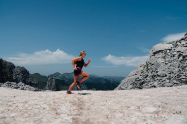 Ekstrem bir sporcu kadın karlı arazide dağlarda gökyüzünde uçuyor. Bir gökyüzü koşucusu dağcılık yapıyor ve vahşi doğada koşuyor. Arka planda bulutlu bir gökyüzü ve dağlar var..