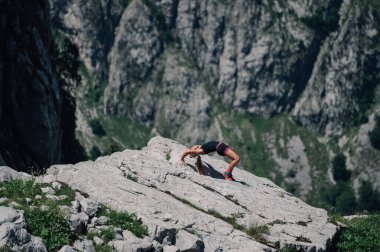 Dişi bir gökdelen kayalık arazide dağlarda esneme egzersizleri yapıyor. Formda bir sporcu kadın dağlarda spor yapıyor ve vahşi doğada pratik yapıyor. Kadın dağdaki uçurumda eğitim görüyor.