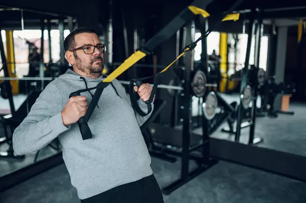 Trx süspansiyon kullanırken orta yaşlı aşırı kilolu adam spor salonunda antrenman yapıyor. Kilo vermeye çalışan şişman bir adam. Tombul adam spor salonunda iyi sonuçlar alıyor..
