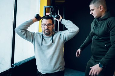 Obez orta yaşlı adamlar spor salonunda kişisel eğitmenin yardımıyla antrenman yapıyor ve kol egzersizleri için çapraz makine kullanıyor. Çapraz antrenman. Sağlıklı yaşam tarzı kavramı.