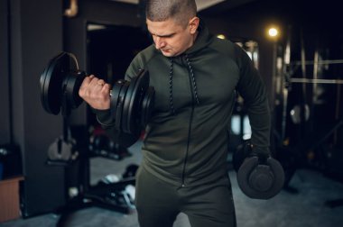 Spor salonunda antrenman yapan ve dambılları kaldıran atletik bir genç. Pazılarını şişiriyor. Sağlıklı yaşam tarzı kavramı.