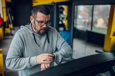 Spor salonunda antrenman yapan şişman, orta yaşlı bir adam koşu bandında koşarken ve akıllı bir şekilde kalp atış hızını kontrol ederken. Sağlıklı yaşam tarzı kavramı.