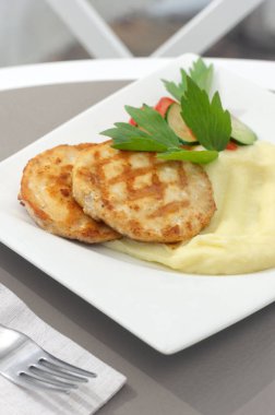 İki çıtır et ya da altın ekmekli tavuk pirzolası, yanında patates püresi, taze dereotu, beyaz seramik tabakta vişneli domates. Bir restoranın arka planında