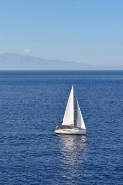 Arka planda kıyı şeridi olan sakin bir denizde, beyaz yelkenli bir yat.