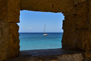 Rhodes, Greece - May 2022: Sailing boat on the sea near the harbour with view framed by a hole in the old town walls.