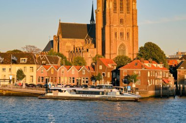 Dordrecht, Netherlands - August 2022: Small fast ferry operated by Blue Amigo docked on the waterfront of the city. In the background is Dordrecht Minster or the Church of our Lady at sunset.