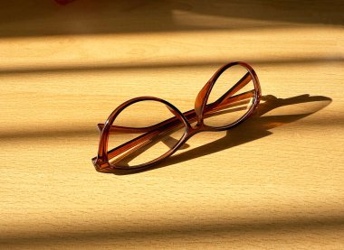 Pair of brown glasses with brown frames on a desk with sunlight sining through a window with blinds.  clipart