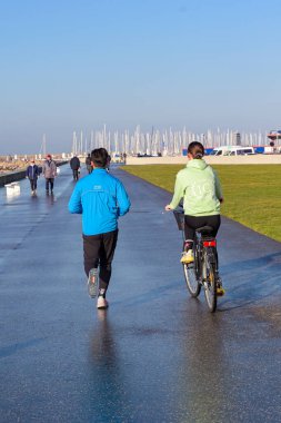 Le Havre, France - 16 January 2025: Person jogging on the promenade of Le Havre with a person riding a bike alongside, clipart