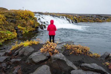 Grafarlandsfoss 'un fotoğrafını çeken kırmızı yağmurluklu kadın Kuzey İzlanda dağlarında renkli sonbahar bitkilerinin arasına düştü..