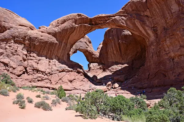 stock image Distant tourists under Double Arch in Arches National Park near Moab, Utah on clear sunny summer morning.