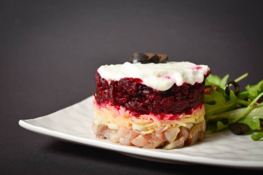 Side view of Soviet layered fish salad Herring under fur coat. Portion with marinated herring, onion, grated potato, beetroot, mayonnaise on top with lettuce olive salad. Dark copy space background
