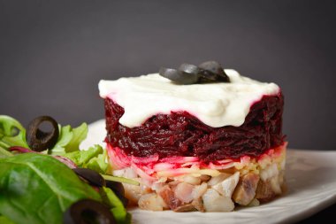 Top view of traditional Russian salad Herring under a fur coat served in culinary ring on white plate. Lettuce leaves and black olives near salad.