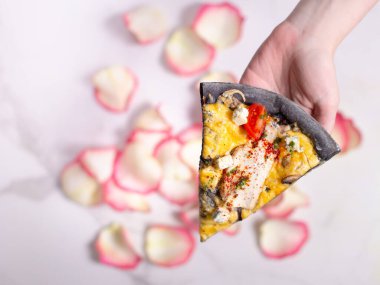 Directly above of black dough pizza slice on marble background with rose petals. Holding pizza in left hand. Romantic scene with food. Pizza with chicken, blue cheese, mushrooms and cherry tomatoes
