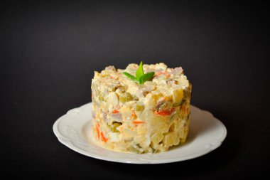 Olivier 'in beyaz tabakta servis ettiği geleneksel Rus salatasına yakın. Patates ve havuç küplü salata, yeşil bezelye, salatalık turşusu, koyu arkaplanda taze soğanlı yumurta.