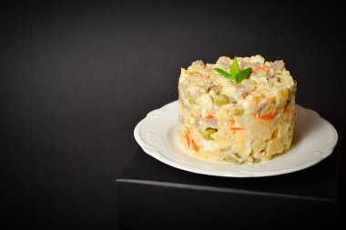Side view of salad Olivier served in culinary ring with greens on top. Dish located on a black box. Copy space image with space for text with dark background. Ukrainian Salad with boiled vegetables