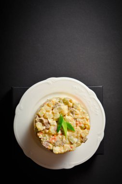 Traditional Soviet salad Olivier served in glass bowl located on black box. Portion of salad with potato and carrot cubes, green peas, pickled cucumbers, eggs, spring onion on top. Dark background.