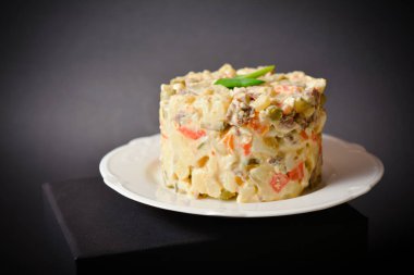 Traditional Soviet salad Olivier served in glass bowl located on black box. Portion of salad with potato and carrot cubes, green peas, pickled cucumbers, eggs, spring onion on top. Dark background.