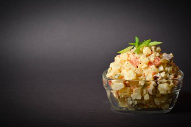 Close up to traditional Russian Olivier salad. Soviet dish served in glass bowl on black background. Salad with green peas, potato, carrot, egg and meat cubes with mayonnaise sauce dressing