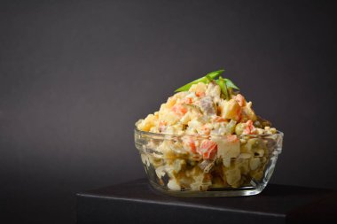 Side view of Traditional Russian salad Olivier served in glass bowl located on box. Salad with potato and carrot cubes, green peas, pickled cucumbers, eggs on Dark background