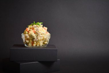 Traditional Russian salad Olivier served in glass bowl located on black box. Salad with potato and carrot cubes, green peas, pickled cucumbers, eggs on Dark background