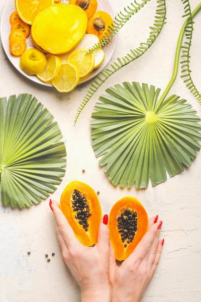 Female hands holding halved papaya fruit on white table with tropical leaves and plate with yellow sliced fruits, top view. Summer food. Healthy eating. Breakfast fruits plate