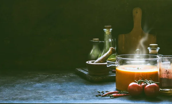 Hot tomato soup with steam in glass pot on rustic table at dark wall background with cooking ingredients. Kitchen scene , still life. Homemade healthy cooking. Vegetarian food.