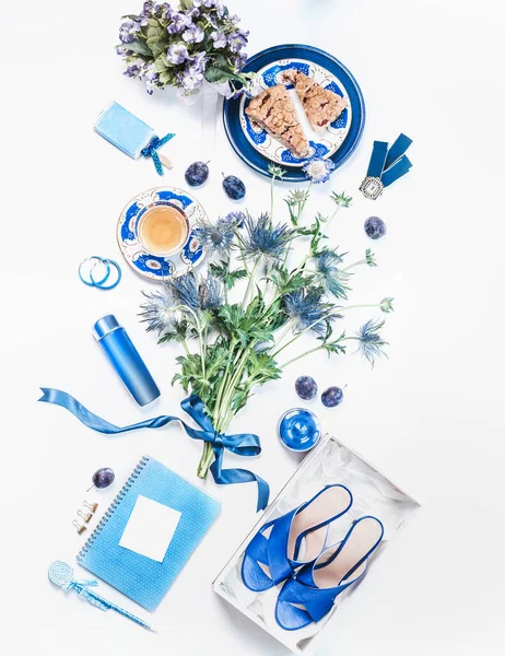Trendy feminine flat lay lifestyle concept. New blue sandals in box, cosmetic products with mock up, flowers bunch, cup of tea with cake pieces, jewelry and notebooks with copy space. Top view. Layout