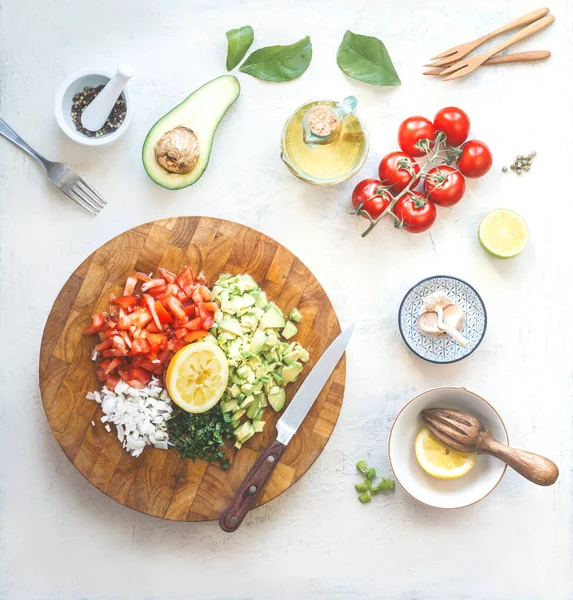 Chopped ingredients for avocado salsa, guacamole  or salad on cutting board. Fresh cut avocado, tomatoes , lemon, lime , olives oil, garlic and chili a on white kitchen table. Top view