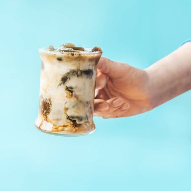 Sütlü buzlu kahve. Mavi arka planda cam bardakta kadın eli. Kapatın. Yaz içeceği konseptini tazelemek