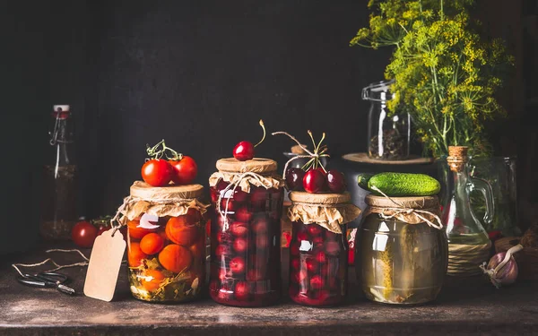 Preserved and fermented  food in glass jars - pickles, jam, compote with tomatoes, cucumber and cherries on dark rustic background.  Fermented food. Autumn canning. Conservation of farm harvest