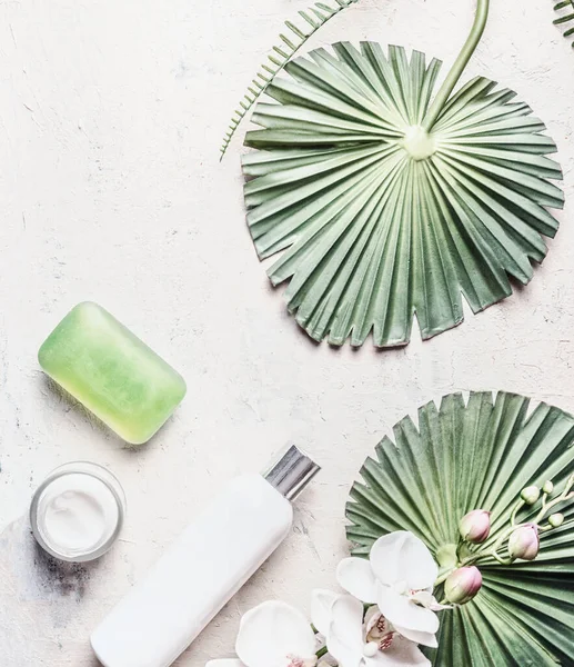 Natural skin care products: bottles, soap and cream on light background with tropical leaves and flowers, top view. Flat lay. Beauty layout. Modern cosmetics. Mock up. Copy space for your design