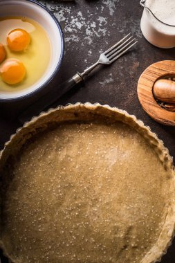 Tart dough in baking form on dark rustic table background with kitchen utensils, top view. Copy space
