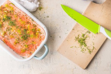 Raw salmon fillet with dill in white casserole on light background with cutting board and kitchen knife, top view. Healthy seafood concept. Diet food. Omega 3