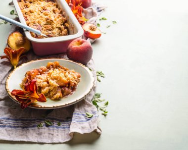 Tasty peach crumble dessert in plate on light background with baking pan and fresh peaches . Copy space
