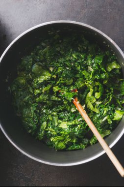 Close up of chopped stewed spinach in cooking pan with wooden spoon on rustic kitchen table. Top view. Vegetarian healthy eating concept.