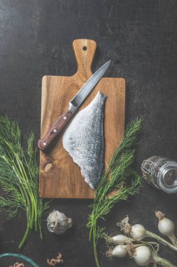 Dorado filetosu mutfak bıçağıyla kesme tahtasında ve koyu mutfak masasında bir demet dereotu ve lezzetli balık pişirme malzemeleri ile birlikte. Yukarıdan görüntüle